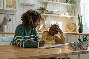 Homeschooling. African American mother sitting beside child son and helping him with homework. Upset schoolboy asking mom to help with math while studying at home. Parental support to learning