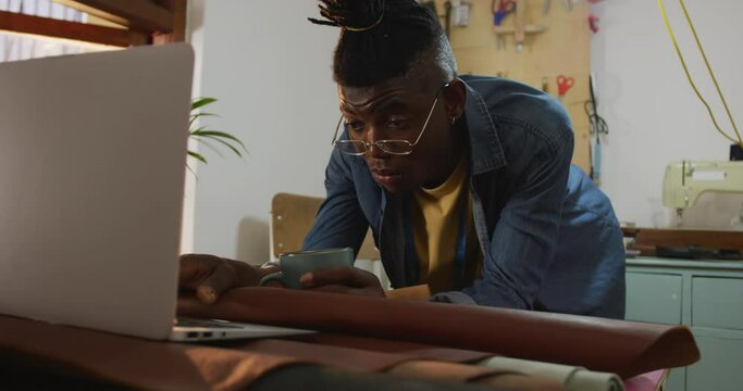 African american craftsman wearing glasses drinking coffee and using laptop in leather workshop