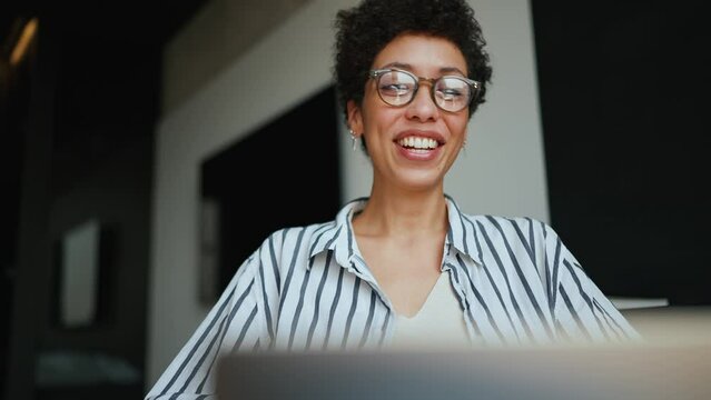 Laughing Hispanic curly haired woman wearing eyeglasses talking by video call on laptop at home