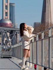 Beautiful young woman with black long hair in white skirt posing eyes closed with Shanghai bund buildings background in sunny day. Emotions, people, beauty, travel and lifestyle concept.