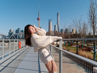 Beautiful young woman with black long hair in white skirt posing eyes closed with Shanghai bund buildings background in sunny day. Emotions, people, beauty, travel and lifestyle concept.