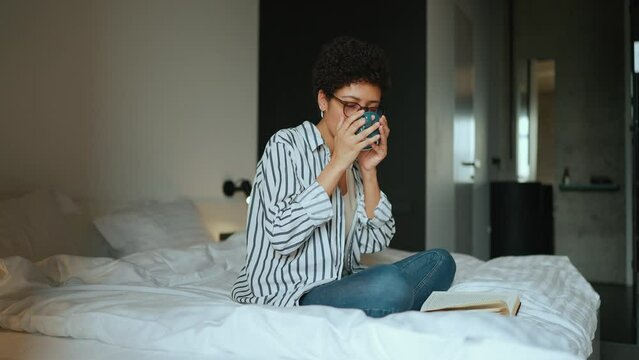Happy Hispanic curly haired woman reading a book and drinking tea on the bed at home