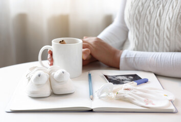 A pregnant woman is waiting leisurely with a teacup, notebooks and baby items are placed in front