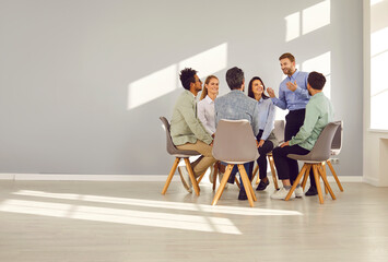Professional business coach and team mentor meeting with group of people. Happy man talking with team of young diverse men and women sitting in circle on chairs in office with grey copy space wall - obrazy, fototapety, plakaty