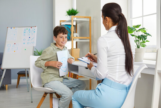Cheerful relaxed preteen schoolboy talks about positive emotions with school psychologist. Child shows sheet with positive emoticon to female psychologist who is taking notes on clipboard.