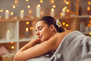 Obraz na płótnie Canvas Young lady getting beauty treatments at a modern spa salon. Happy, relaxed woman with closed eyes lying on the bed, with beautiful candle lights gleaming in the background. Spa, female beauty concept