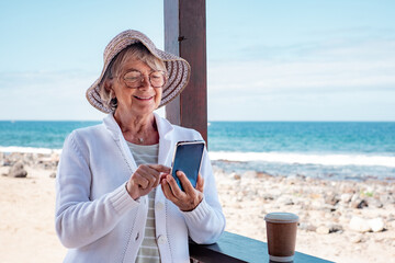 Smiling senior woman wearing hat and eyeglasses standing at the beach using mobile phone typing message, horizon over sea and blue sky