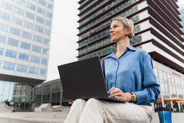 Blond business woman working on laptop sitting outdoors office building