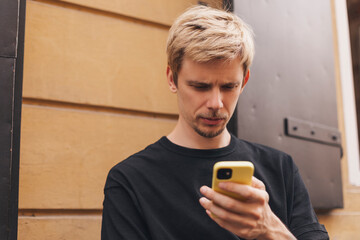 Fototapeta premium Calm relaxed male freelancer in casual clothes focusing on screen, read news, messaging and using smartphone with interest while sitting alone at table on terrace of modern coffee shop.