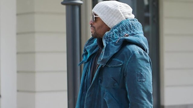 Authentic Portrait of handsome African American man walking in city, wearing stylish outfit parka coat, hoodie, knitted hat and sunglasses. street style fashion male model