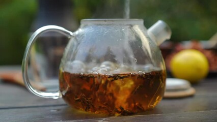 Transparent glass teapot with healthful hot drink and water pouring inside. Close-up green tea outdoors on calm autumn fall day