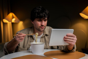 Man eats Chinese noodles while browsing and using a tablet at the same time. The concept of smartphone addiction. 