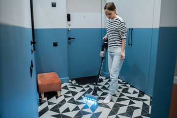 Young woman vacuums the floor in the hallway next to the front door. 