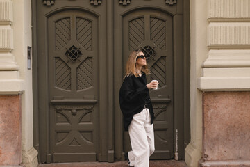 Happy beautiful stylish woman having good fashion white jeans pants, black shirt walking by street, make step and holding coffee or tea in cup takeaway, good mood. Girl look happy, go away from house.