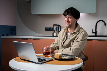 Young freelance guy is watching a movie on a laptop while sitting at a table in the kitchen. A man eats a piece of cake on a saucer, drinks tea 