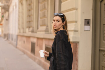 Calm beautiful stylish woman having good fashion clothes walking on street and holding some coffee in cup takeaway with good summer mood. Girl look happy, turn around, turn back or spinning.