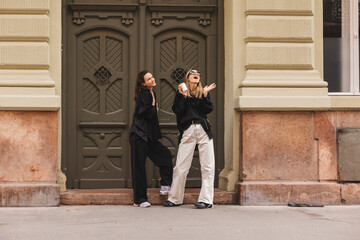 Two happy smiling women standing near door outdoor on the street and look at side with wow emotions. One woman hold cup of coffee, open mouth, two surprised girls sisters tourists look oh it so sweet.