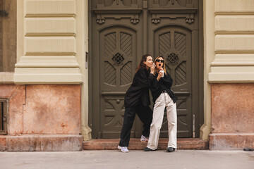 Two pretty cuter sisters shares secrets, gossip. Surprise face, emotions, Best friends wearing stylish outfit, sunglasses, shirts, pants. Two girls standing near doors on the street. Women friendship.