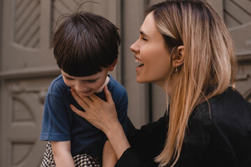 Happy mom tell secret or joke on ear her son. They look happy and laughing. Mother and children near door outside, walking on the street. Happy family, parent, childhood concept.