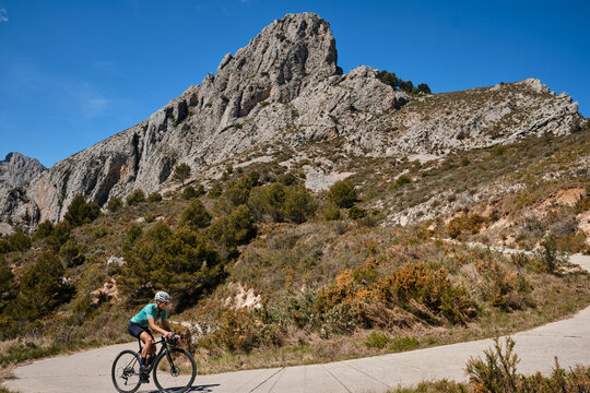 A female cyclist climbing a mountain road on a gravel bike, wearing a cycling kit and helmet.The cyclist riding between mountains, creating a beautiful and motivational image of an athlete.Spain