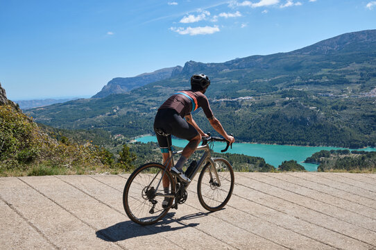 Man cyclist in helmet and cycling kit riding through mountainous roads. Cyclist on gravel bike with scenic views of turquoise lake in Spain.Gravel riding through the mountains.Guadalest Reservoir.