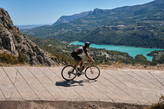 Man cyclist in helmet and cycling kit riding through mountainous roads. Cyclist on gravel bike with scenic views of turquoise lake in Spain.Gravel riding through the mountains.Guadalest Reservoir.