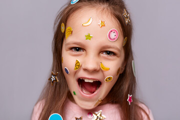 Closeup image of funny crazy little brown haired little girl covered with stickers posing isolated over gray background, looking at camera, screaming loud.