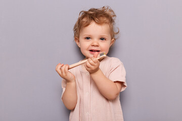 Indoor shot of cute blonde wavy haired toddler baby girl brushing her teeth, looking at camera, playing with tooth, standing isolated over gray background.