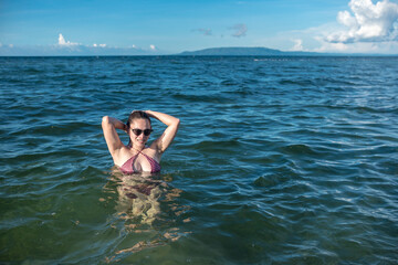 Fototapeta na wymiar A sexy asian woman in a bikini and wearing shades wading in chest deep waters during a sunny day at the beach