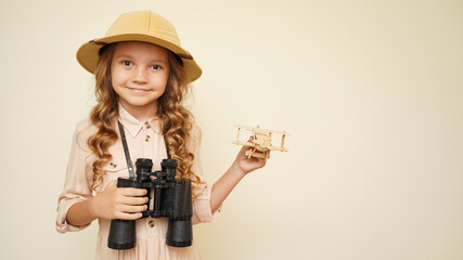 Unique child. The child dreams of traveling, flying, new discoveries. The kid wants to be a traveler, discoverer, explorer. A little girl holds a wooden airplane and binoculars, wearing a safari hat. © AlyaRo