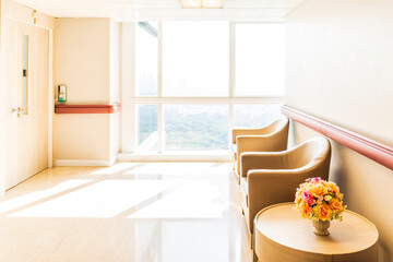 Warm tone of chairs or sofa in front of private room in modern hospital corridor or walkway with sunray background.Glasses window shows park landscape.Clean floor and reflection with roses decorated.