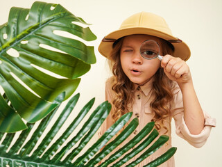 Child discoverer. Traveler child. Young explorer. A little girl in a safari hat with a magnifying glass looks carefully through the green palm leaves. Concept: search for adventure and treasure.