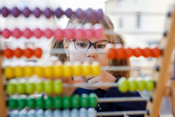 Little preschool girl playing with educational wooden rainbow toy counter abacus. Healthy happy child with glasses learning to count and colors, indoors on sunny day.