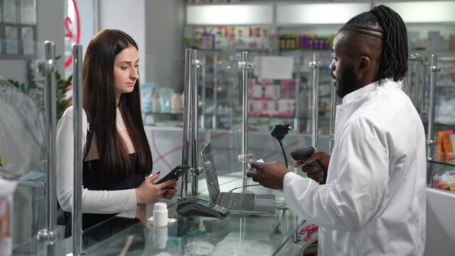 Witness the checkout process at the counter of a modern drugstore as male African American pharmacist scans medicine and young female customer pays contactless with her smartphone. 4K high-quality.