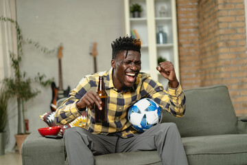 Obraz premium Young African man watching sport match on television and supporting his favorite team. Happy man enjoying victory, watching sports competition on tv.