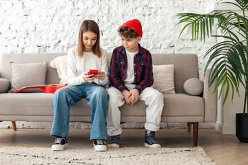 Teenagers a boy and a girl are sitting on a sofa in a loft-style bright room, a girl opens a red box presented by a boy. Gift for birthday, Valentine's day, Christmas. Teenage relationships