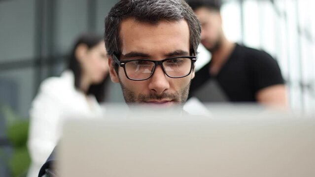 Portrait of handsome bearded man wearing glasses working with laptop in office late, his face lit up by screen