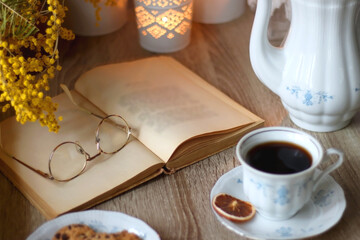 Cup of tea or coffee, plate of cookies, cup of blueberries, plate of chocolate, glass of juice, book, reading glasses, teapot, flowers and lit candles on the table. Brunch or afternoon tea concept.