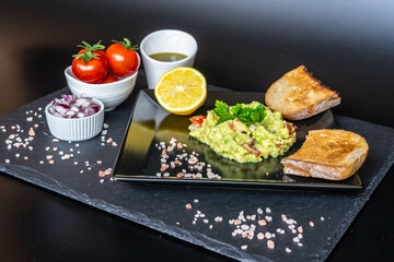 Homemade guacamole with red onion, cherry tomatoes, olive oil, pink salt and lemon juice, accompanied by toast . Home cooking, eating healthy, vegan food