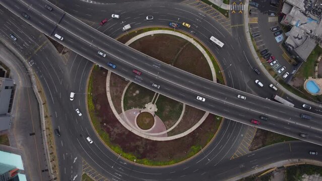 Cars drive on an overpass, a street called "Javier Prado" above a roundabout called "Ovalo Monito Huascar". Drone slowly flies down while rotating left. Located in Lima, Peru.