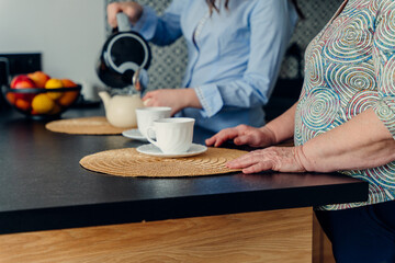 Unrecognizable teenage granddaughter and her wonderful grandmother drink tea in a modern kitchen interior. Focus on older woman hands.