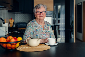 Portrait of an elderly old woman in glasses sitting in the kitchen and drinking tea alone and looking positively at the camera.