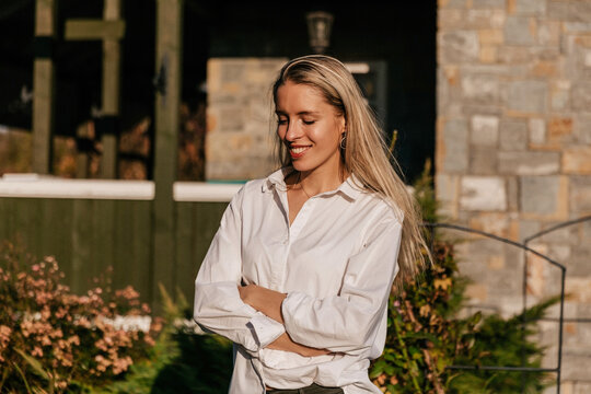 Close up outdoor photo of smiling happy girl with wonderful smile and long blond hair wearing white shirt is enjoying time at home garden in sunlight in summer evening