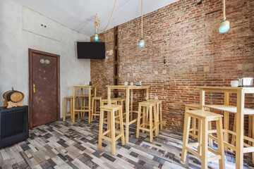 An empty bar with a rope lamp, a ceiling fan, raw brick walls and high white wooden tables with matching stools, a wooden mosaic floor and a television