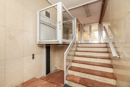 Access portal to a residential building with stairs and tiled floors of brown marble with veins, walls in cream marble and an elevator for people with reduced mobility