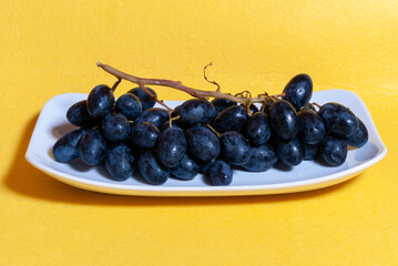 Uvas negras en plato blanco, con fondo amarillo con textura rugosa.