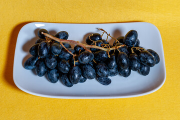 Uvas negras en plato blanco, con fondo amarillo con textura rugosa.