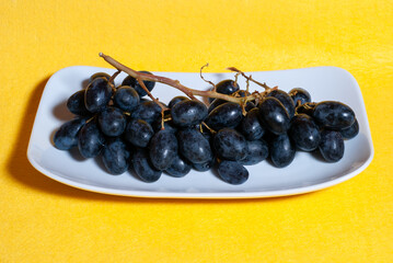 Uvas negras en plato blanco, con fondo amarillo con textura rugosa.
