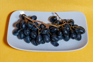 Uvas negras en plato blanco, con fondo amarillo con textura rugosa.