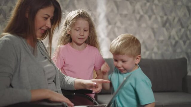 Mother and sister watch toddler pushing buttons on keyboard of laptop. Woman with paralysis sits in wheelchair using device with kids closeup